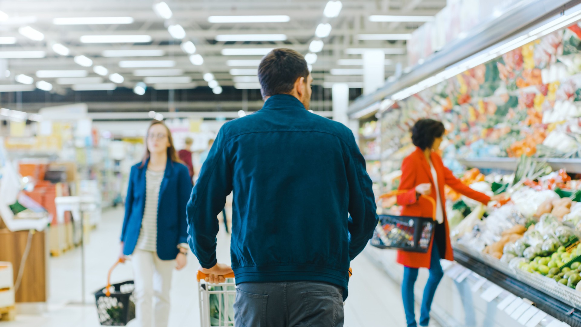 At the Supermarket: Man Pushing Shopping Cart Through Fresh Produce Section of the Store. Store with Many Customers Shopping. Following Back View Shot.